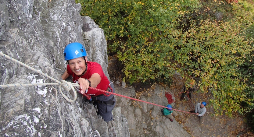 A person wearing safety gear is secured by ropes as they look up and smile while rock climbing.